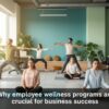 The image shows a group of employees participating in a yoga session in a modern office space with a vibrant green background. Several individuals are performing different yoga poses, while others are sitting in a relaxed position with their eyes closed, engaging in mindfulness. The room is filled with plants and cozy cushions, creating a calming atmosphere. The text "Why employee wellness programs are crucial for business success" is displayed at the bottom of the image.